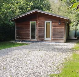 A wooden cabin with a gravel driveway and surrounded by trees at 1 Streamside in Lanreath