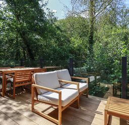 An outdoor wooden deck with chairs a table and glass railing surrounded by trees at 1 Streamside in Lanreath