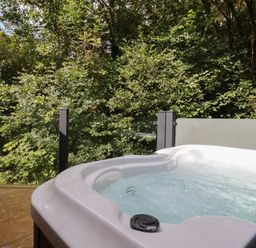 A hot tub on a wooden deck with glass panel fencing and dense green trees behind at 1 Streamside in Lanreath