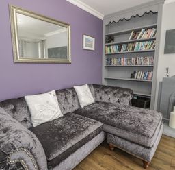 A living room with a purple wall, a gray velvet sofa with white pillows, a mirror, a bookshelf with books and DVDs, and a gray tiled fireplace at Tan Y Fron in Dyffryn Ardudwy