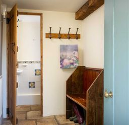 A hallway with an open door revealing a bathroom with tiled walls a wooden bench with boots underneath and a wall-mounted coat rack at Hope Cottage in Longborough