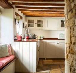 A kitchen with wooden beams stone wall window seat with cushions and cabinets at Hope Cottage in Longborough