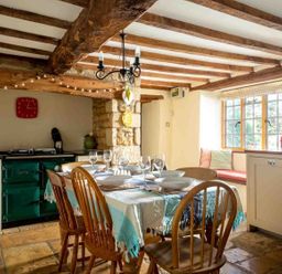 A dining area with a table set with dishes and glasses wooden chairs a window bench with cushions a green stove and wooden ceiling beams at Hope Cottage in Longborough