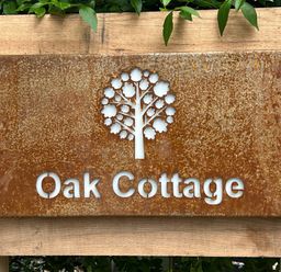 A rusted metal sign with a tree cutout and Oak Cottage text mounted on wood with green leaves at Oak Cottage in Somerleyton