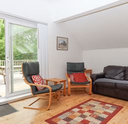 A sitting area with two wooden armchairs with cushions a leather sofa a small wooden table a patterned rug and sliding glass doors leading to a wooden deck at Crystals in Hatherleigh