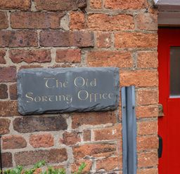 A brick wall with a black slate sign reading The Old Sorting Office next to a red door at The Old Sorting Office in Hawarden