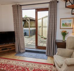 A living room with a TV on a wooden stand a beige armchair a side table with flowers and a lamp and glass doors leading to a patio at The Old Sorting Office in Hawarden