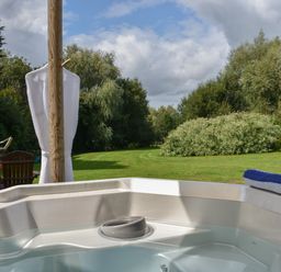 An outdoor hot tub with water and a towel on the edge overlooking a grassy yard with trees in the background at Burr 1 Shepherds Hut in Barnstaple