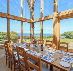 A dining area with a wooden table and chairs set with plates glasses and crab-themed placemats in a room with large windows overlooking a garden and ocean at Pen y Graig in Aberporth