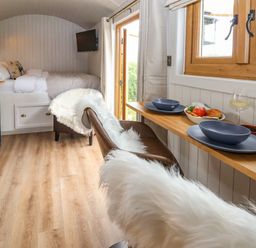 A small room with a bed a wall-mounted TV a kitchenette with blue cabinets a wooden dining counter with two plates and glasses and two chairs with white fur throws at Cariad Mawr in Gwaenysgor near Prestatyn