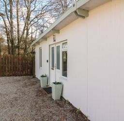 The exterior entrance of a white building with two windows a door two tall planters and a wooden fence behind trees at Caldwell Cottage in Lanark