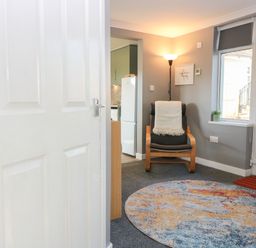 A sitting area with a wooden armchair with a blanket a round colorful rug a floor lamp and a window at Caldwell Cottage in Lanark