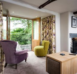 A living room with a purple armchair a green chair a wooden coffee table floral curtains and a fireplace at The Cosy Peacock in Troutbeck