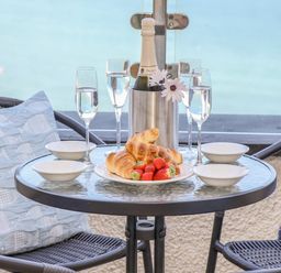 A round glass table with croissants strawberries four bowls four glasses of water and a bottle in an ice bucket with flowers on a balcony with two chairs and cushions at Mariners Watch in Carbis Bay