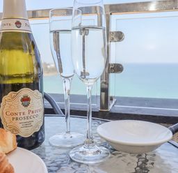 A bottle of Prosecco with two filled glasses on a glass table with a croissant and a white dish on a balcony overlooking the sea at Mariners Watch in Carbis Bay