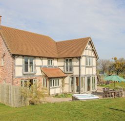 An exterior view of a two-story house with a patio area including a table with chairs and an umbrella and a hot tub in a grassy yard at Cuckoo Pen Cottage Eastham near Tenbury Wells