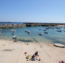 A beach with people sitting on sand and boats anchored in a harbor at The Net Loft in Mousehole