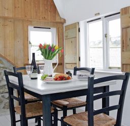 A dining area with a white table black chairs a plate of food a bottle of wine glasses and flowers at The Net Loft in Mousehole