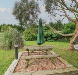 An outdoor area with a wooden picnic table and bench under a closed green umbrella near a tree with a hanging chair at The Stables in Ruthvoes near St Columb Major