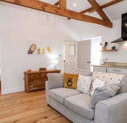 A living room with a gray sofa and kitchen area with wooden beams on the ceiling at Pear Tree Cottage in St Mellion near St Dominick
