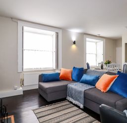 A living room with a gray sectional sofa with blue and orange cushions next to two windows with white shutters and a small dining table in the background at Carew House Cottage in St Day