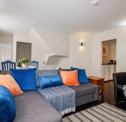 A living room with a grey sectional sofa with blue and orange cushions and a dining table with blue chairs at Carew House Cottage in St Day