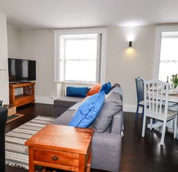 A living room with a gray sofa with blue and orange pillows a wooden side table a dining table with chairs and flowers on the table at Carew House Cottage in St Day