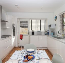 A kitchen with white cabinets a dining table set with plates and glasses a microwave on the wall and windows with blinds at Sandy Toes Paignton