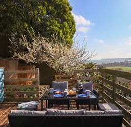 An outdoor wooden deck with a table set for breakfast and cushioned chairs overlooking a coastal view at Sandy Toes in Paignton