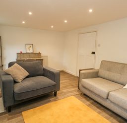 A living room with a gray armchair, gray sofa with a crab pillow, wooden floor, yellow rug, shelving unit, and a desk at Half Pint Cottage in Beaminster