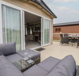 An outdoor deck with gray cushioned seating and a glass table with wine glasses adjacent to a dining table with chairs at Whispering Willows in Cayton Bay