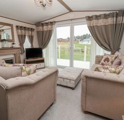 A living room with two beige sofas a patterned ottoman a TV on a wooden stand a fireplace with a mirror and windows with curtains at Whispering Willows in Cayton Bay