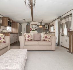 A living room with beige sofas and a patterned ottoman next to a kitchen and fireplace at Whispering Willows in Cayton Bay