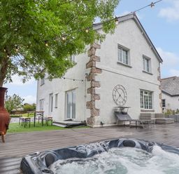 An outdoor area with a bubbling hot tub under a tree string lights bench table two lounge chairs a large clock on the wall and a white house at The Manor House Skerry Vore Bryn Pydew near Llandudno Junction