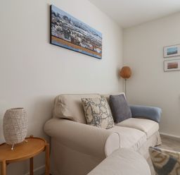 A living room with a beige sofa cushions a side table with a woven lamp floor lamp and coastal paintings on the walls at 16 Church Street in Lyme Regis