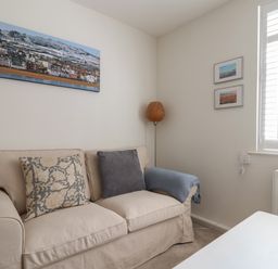 A beige sofa with cushions and a blue throw near a corner with a wicker floor lamp and framed pictures beside a window with white shutters at 16 Church Street in Lyme Regis