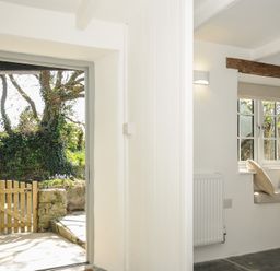 An open door leading to a garden with a wooden gate and a window seat with cushions inside a white room at Sunrise Cottage in St Kew Highway near Wadebridge