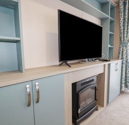 A TV on a wooden cabinet above an electric fireplace with built in shelves and blue cabinets near a window with patterned curtains at The Mermaid A04 in Par