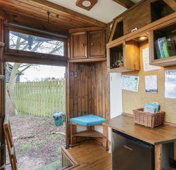 A small wooden room with a door opening to a fenced garden a mini fridge a wooden desk and cushioned corner seats at Daisy Cow Shed 1 Easton near Campsea Ashe