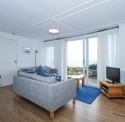 A living room with gray sofas blue cushions a small wooden TV stand with a flat screen TV a blue rug and sliding glass doors leading to a garden at Ael Y Don in Llandanwg