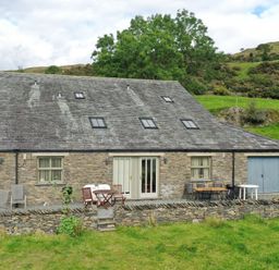 Stone building with slate roof chairs and tables on stone patio with grassy hill in background at Ghyll Bank Barn in Staveley near Windermere