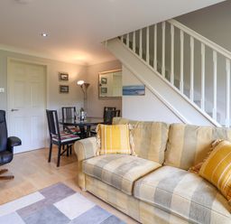 A living room with a striped sofa with yellow pillows a black armchair a round dining table with four chairs and a staircase at 50 Fernhill Heights in Charmouth