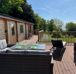 A wooden deck with outdoor seating and dining set next to a wooden cabin surrounded by trees at Northwood Lodge in Llanbedrog