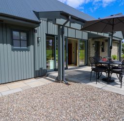 An outdoor patio area with a round table and four chairs under an umbrella next to a building with sliding glass doors at Bramble Cottage in Wester Essendy near Dunkeld