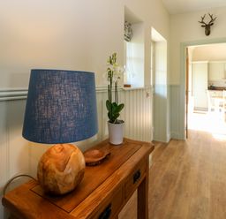 A hallway with a wooden table holding a lamp and a potted orchid with a doorway leading to a kitchen at Bramble Cottage Wester Essendy near Dunkeld