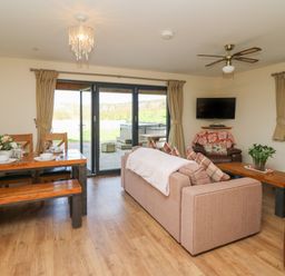 An open living room and dining area with a sofa, wooden dining table with benches, armchair, wall-mounted TV, and large sliding glass doors at Caban Dulas in Rhayader