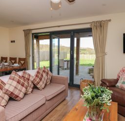 A living room with a brown sofa with plaid cushions a brown leather armchair a wall-mounted TV and dining table with chairs near glass doors at Caban Dulas in Rhayader