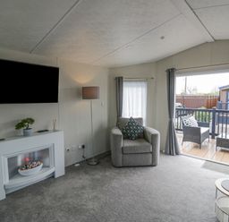 A living room with a wall-mounted TV a white electric fireplace an armchair and a floor lamp next to sliding glass doors leading to an outdoor seating area at Long Acres Lodge in Evesham