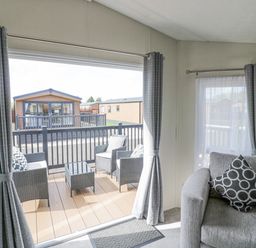 A living area with gray armchair and patterned cushions next to a sliding door leading to a deck with outdoor chairs and table at Long Acres Lodge in Evesham