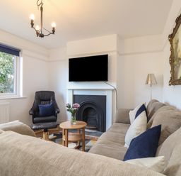A living room with a beige sofa and blue cushions a black armchair a small round wooden table with flowers a wall-mounted TV and a fireplace at The Heights in Truro